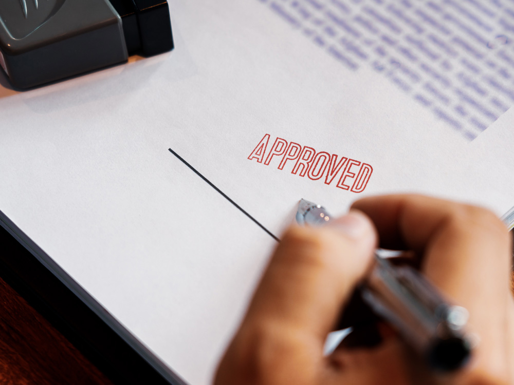 person signing document with the word "approved" stamped on the paper in red ink